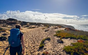 Hiker with blue backpack walking on sandy coastal path at Praia do Brejo Largo, Portugal. Ocean and dramatic clouds visible under sunny sky.