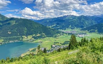 Panoramablick von der Schmittenhöhe über den Zeller See, grünes Tal und Bergketten im Pinzgau unter blauem Himmel mit weißen Wolken.