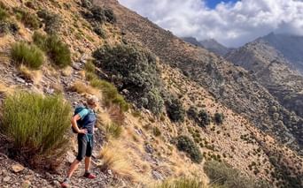 Wanderin mit Rucksack auf schmalem Bergpfad in den Alpujarras, Andalusien. Steile felsige Hänge mit Grasbüscheln und dramatische Berggipfel unter bewölktem Himmel.