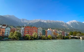 Row of colorful historic houses along the Inn River in Innsbruck with snow-capped Alpine mountains in the background under clear blue sky.
