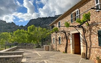 Stone building Refugi Tossals Verds with patterned courtyard, vine-covered entrance, and mountain backdrop on Mallorca's GR221 trail.