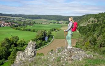Hiker with red backpack standing on rocky outcrop overlooking green valley in Altmühltal with villages, fields, and forested hills.