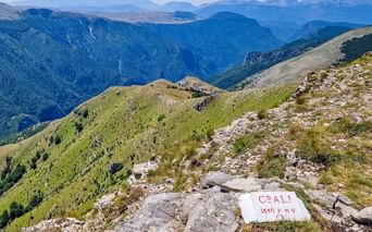 White summit marker reading 'OBALJ 1896 m n.v.' on rocky mountain peak with panoramic view of Bjelasnica mountain ranges in Bosnia.