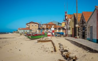 Bunte Fischerboote am Sandstrand in Vila Chã mit traditionellen Fischerhäusern mit orangefarbenen Ziegeldächern unter blauem Himmel.
