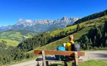 Two hikers with backpacks sit on a wooden bench overlooking green alpine meadows and the rocky peaks of the Kitzbühel Alps under a blue sky.