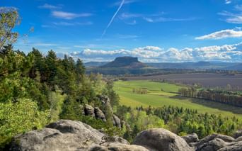 Blick von Rauenstein-Felsformationen über grüne Felder und Wälder zum markanten Tafelberg Lilienstein unter blauem Himmel mit Wolken.