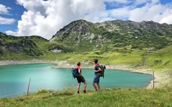 Zwei Wanderer mit Rucksäcken bewundern den türkisfarbenen Formarinsee, umgeben von grünen Almwiesen und felsigen Bergen unter blauem Himmel.