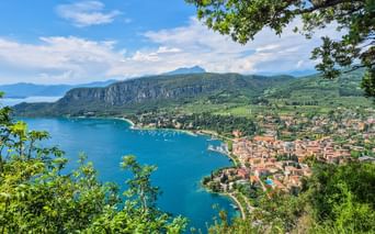 Panoramic view of a town along Lake Garda's turquoise bay, framed by green foliage. Mountains rise behind the settlement with boats dotting the water.