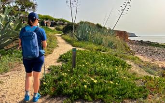 Wanderer mit blauem Rucksack auf sandigem Küstenweg der Rota Vicentina. Agaven und Meeresklippen unter bewölktem Himmel sichtbar.