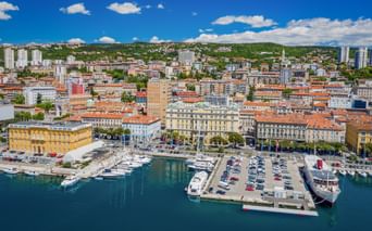 Aerial view of Rijeka harbor in Kvarner Bay with moored yachts, parking area, and historic yellow buildings along the waterfront under blue sky.