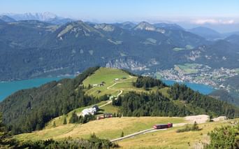 Panoramic view from Schafberg mountain showing green alpine meadows, a turquoise lake, and mountain ranges in Salzkammergut, Austria.