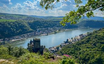 Aerial view of Burg Katz castle overlooking the Rhine River and St. Goarshausen town, surrounded by green hills under blue sky with clouds.