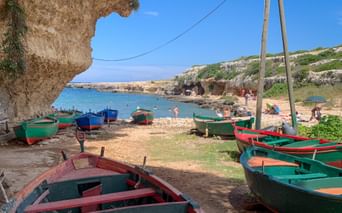 Bunte Holzboote am Sandstrand in Polignano, Apulien. Felsige Klippen rahmen die Bucht mit türkisfarbenem Wasser und Strandbesuchern ein.