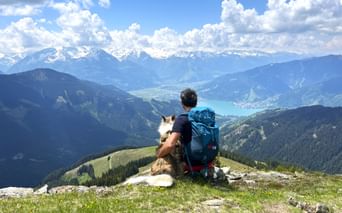 Wanderer mit Rucksack und Hund auf Berggipfel im Pinzgau mit Blick auf den türkisfarbenen Zeller See umgeben von Alpengipfeln.