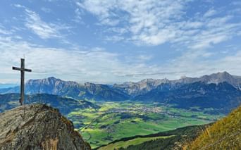 Summit cross on rocky peak overlooking green valley and Kitzbühel Alps mountain range under blue sky with scattered clouds.