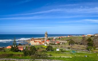 Küstenansicht des Klosters Santa Maria de Oia mit Glockenturm, umgeben von grünen Feldern und rotgedeckten Gebäuden mit Blick auf den Atlantik.