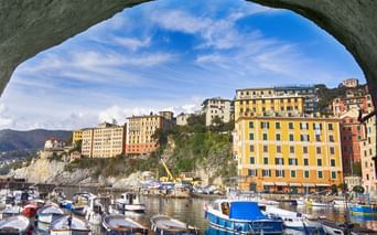 Blick auf den Hafen von Camogli durch einen Steinbogen mit bunten Gebäuden am Hang, Fischerbooten im Wasser und blauem Himmel.