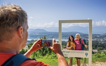 Man taking photo of two women posing at Velden photo frame with lake and mountains in background. Clear blue sky over Carinthian landscape.