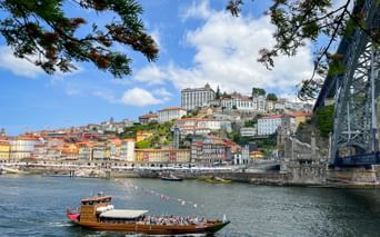 Traditional wooden boat on the Douro River with Porto's colorful hillside buildings and bridge visible. Blue sky with white clouds above the cityscape.