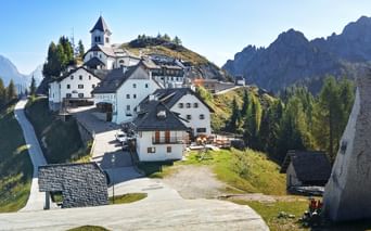 Monte Lussari mountain village with white church and traditional buildings on a hillside, surrounded by forests and rocky peaks under blue sky.