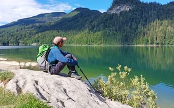 Female hiker with backpack and poles sitting on rock at Schwarzensee lake, Salzkammergut. Forested mountains reflect in calm green water.