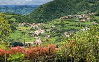 Panoramic view of an Italian hillside village with scattered houses among green rolling hills, vineyards in foreground, mountains in background.