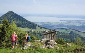 Zwei Wanderer mit Rucksäcken auf einem Bergweg mit Blick auf den Chiemsee. Eine Holzhütte steht auf Felsen, bewaldete Gipfel und Tal darunter.
