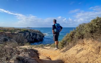 Wanderer mit blauem Rucksack auf sandigem Weg mit Blick auf felsige Klippen und blauen Atlantik bei Porto Covo an der Rota Vicentina.