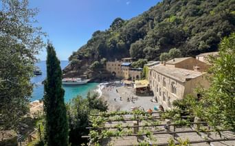 San Fruttuoso bay with historic abbey buildings, pebble beach, and turquoise water. Lush green hillside rises behind the beige stone structures.