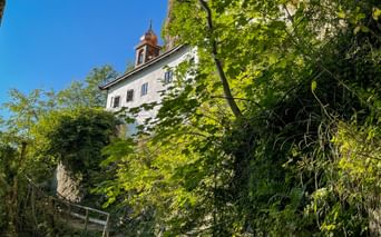 White hermitage building with bell tower perched on steep rocky cliff surrounded by lush green trees under blue sky in Salzburg region.