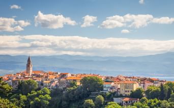 Coastal town of Vrbnik on island Krk with church tower, terracotta roofs, and lush greenery. Mountains and blue sea visible in background.