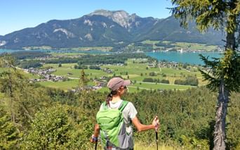 Female hiker with green backpack and trekking poles viewing Wolfgangsee lake in Salzkammergut. Mountains, green valleys, and villages visible below.