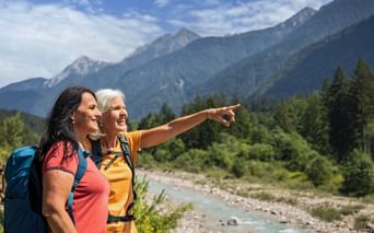 Two women hikers with backpacks admiring mountain view near Villach. One points across a turquoise river toward forested Alpine peaks.