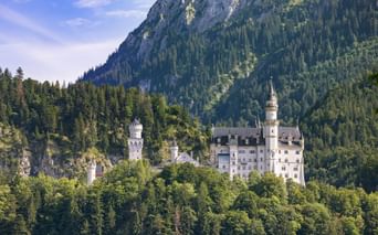 Neuschwanstein Castle nestled among dense forests with dramatic rocky mountain peaks rising behind it under a blue sky.
