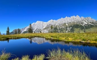Hochkönig mountain range with snow-capped peaks perfectly reflected in the calm waters of Spiegelsee, surrounded by green meadows and spruce trees.