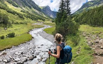 Wanderer mit blauem Rucksack und Stöcken betrachtet Gebirgsbach im grünen Tal. Steile Hänge und Nadelbäume rahmen den Lechweg.