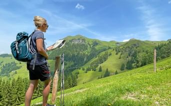 Female hiker with blue backpack reading a map on a green alpine meadow in Kitzbühel, with forested mountains under blue sky.