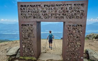 Wanderer mit Trekkingstöcken geht durch das metallene Granattor mit ausgestanztem Text auf einem Berggipfel. Bergketten im Hintergrund unter blauem Himmel.
