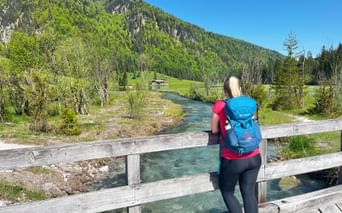 Female hiker with blue backpack on wooden bridge over turquoise river near Pillersee, forested mountains and meadows in background.