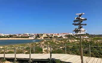 Wooden boardwalk with directional signpost on coastal path overlooking river and Vila Nova de Milfontes town under clear blue sky.