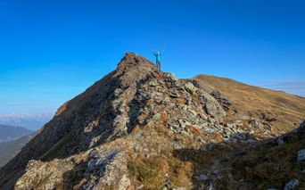 Female hiker with raised arms celebrating on rocky mountain summit in the Kitzbühel Alps under clear blue sky with mountain ranges visible.