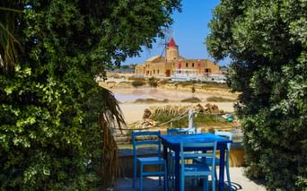 Blick auf Trapani Windmühle durch Gartenterrasse Blaues Outdoor-Essset auf Terrasse umrahmt von grünem Laub, mit Blick auf historische Windmühle und Salzfelder von Trapani, Sizilien.