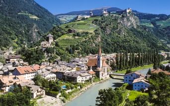 Alpine town of Klausen with church spire, river, and historic castles on hillsides. Mountains and vineyards visible in background under blue sky.