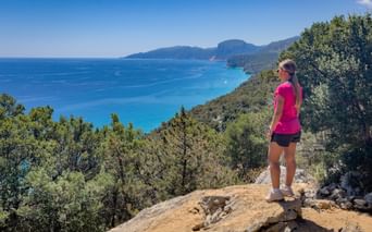 Woman in pink shirt standing on rocky viewpoint overlooking turquoise Kvarner Bay with green hills and distant mountains under blue sky.