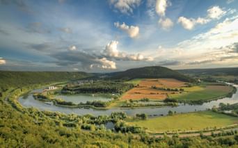 Luftaufnahme des Altmühltals mit einem sich windenden Fluss, der eine Schleife um grüne Felder und Wälder bildet, unter blauem Himmel mit weißen Wolken.