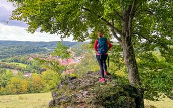 Female hiker with backpack standing on rocky outcrop under large tree, overlooking village of Pappenheim in Altmühltal valley with green hills.