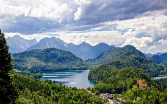 Aerial view of Hohenschwangau village with yellow castle tower, surrounded by forested hills and blue Alpine lakes, with mountain peaks in background.