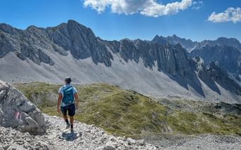 Hiker with backpack walking on rocky mountain path in Karwendel with dramatic jagged peaks and scree slopes under blue sky with clouds.