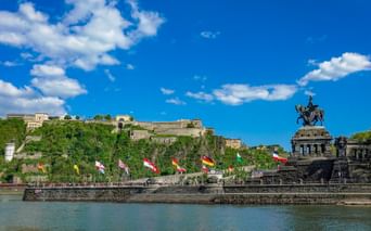 Deutsches Eck Denkmal mit Reiterstatue in Koblenz, bunte Flaggen am Rheinufer, Festung Ehrenbreitstein auf dem Hügel.