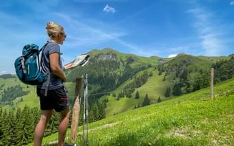Female hiker with blue backpack reading a map on a green alpine meadow in the Kitzbühel Alps, with forested mountains under blue sky.
